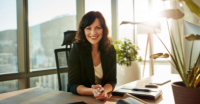 Professional woman smiling warmly at modern desk with plants and sunlight streaming through tall windows overlooking mountains